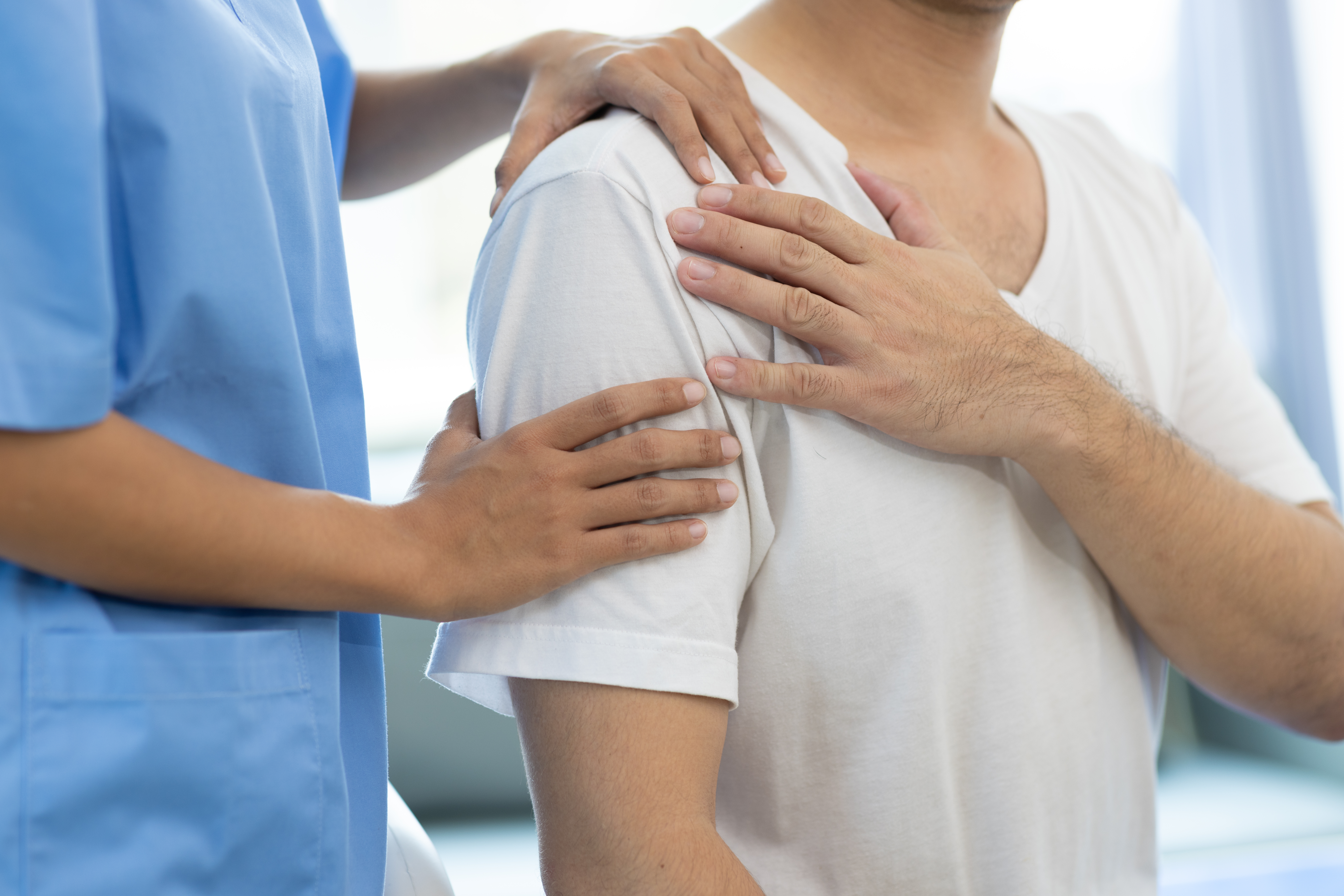Doctor diagnosing man's shoulder pain in hospital examination room. Nurse doing massage and physiotherapy to treat patient's shoulder.